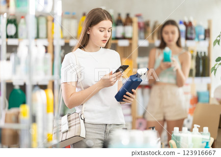 Young female shopper scans QR code on label of spray glass cleaner for windows using her smartphone, while in background shoppers browse products 123916667
