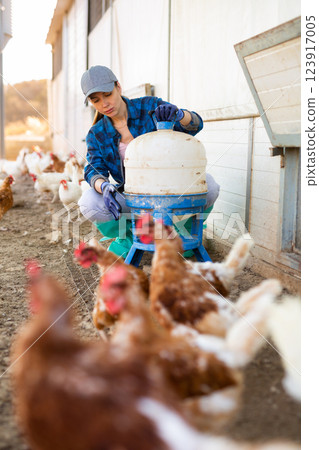 Female farmer holding legged bird drinker while chicken walking on backyard 123917005