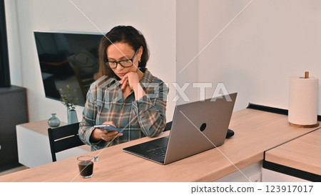 Woman Working Remotely Using Laptop and Smartphone in Cozy Modern Home Office Woman Working Remotely Using Laptop and Smartphone in Cozy Modern Home Office 123917017