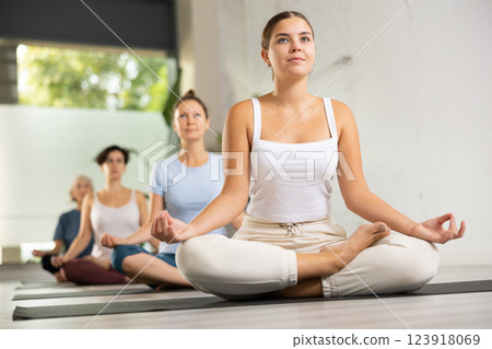Calm young woman sitting in Lotus Pose Padmasana on mat during meditation and group yoga class in fitness studio 123918069
