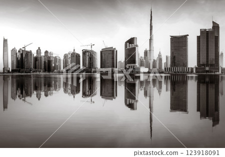 Monochrome city skyline with tall skyscrapers reflecting on calm water under a cloudy sky, Dubai Marina bay UAE 123918091