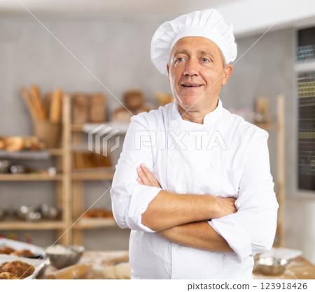 Mature man baker in white coat overall and cap stands in kitchen of bakery 123918426