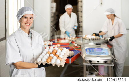 Cheerful Hispanic workwoman of poultry farm holding chicken eggs in cardboard tray 123918524