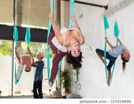 Happy latin woman practicing aerial yoga asana, hanging during group class in gym Happy latin woman practicing aerial yoga asana, hanging during group class in gym 123918699