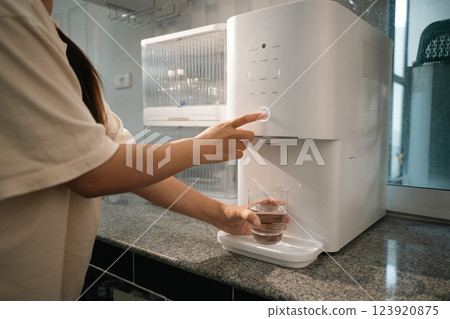 Young woman pressing button on a modern water purifier, filling a glass with fresh water 123920875