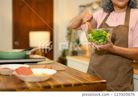 Cropped shot mature woman mixes fresh green salad in a glass bowl while standing in a kitchen 123920876