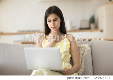 Serious young woman sitting on couch reading email on notebook 123921697