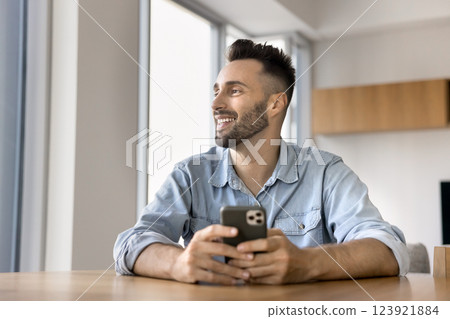 Happy Latin man looking away holding smartphone seated at table 123921884