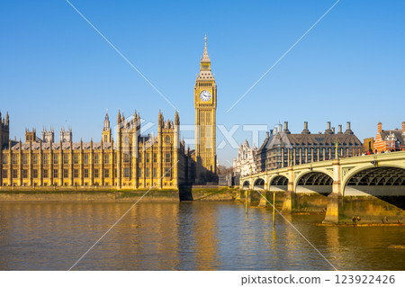 Sunlight reflects on the River Thames as the Houses of Parliament and Big Ben stand proudly in London. Westminster Bridge connects the iconic landmarks, depicting a vibrant day. 123922426