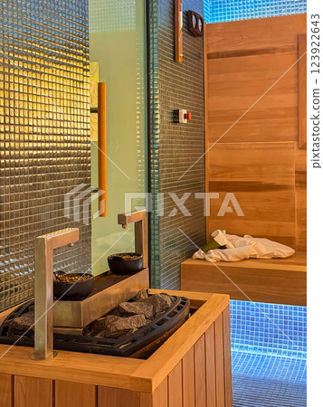 Heater with hot stones in sauna. Modern sauna interior with a wooden heater, walls, glass door, warm lighting. Relaxing wellness space with mosaic tiles and steam stones 123922643