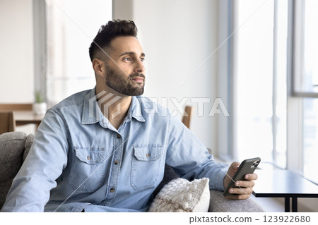 Man sit on couch, holds smartphone, gazing thoughtfully into distance Man sit on couch, holds smartphone, gazing thoughtfully into distance 123922680