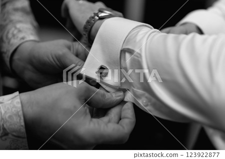 A man fastens a cufflink on the groom's shirt.The groom fastens the cufflinks on his shirt.The groom has a watch on his hand,he is wearing a white shirt.Photography sets up the mood of the celebration 123922877