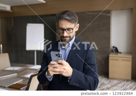 Focused male entrepreneur standing in conference room using his smartphone Focused male entrepreneur standing in conference room using his smartphone 123922984