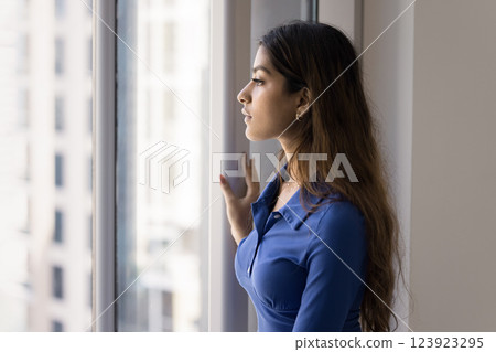 Indian woman standing by window, gazing outside with contemplative expression 123923295