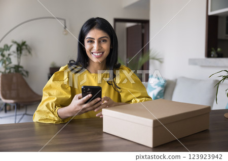 Happy woman sits at table with parcel holding her smartphone 123923942