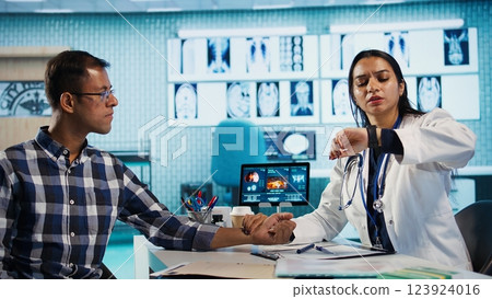 Expert using smartwatch to measure patient heart rate and heartbeat during a medical check up visit. Consultation involves pulse and blood pressure analysis at appointment in office. Camera B. 123924016