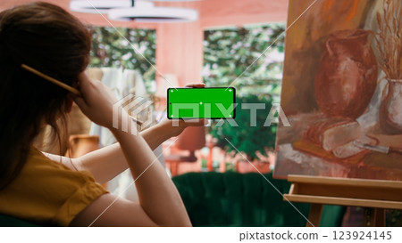 Female artist holding device with green screen in art and craft atelier, admiring rustic artwork painted with watercolor paint. Woman preparing masterpiece for art gallery exhibition. Camera B. 123924145