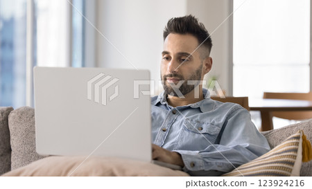 Focused Hispanic man settled down comfortable on couch with laptop Focused Hispanic man settled down comfortable on couch with laptop 123924216