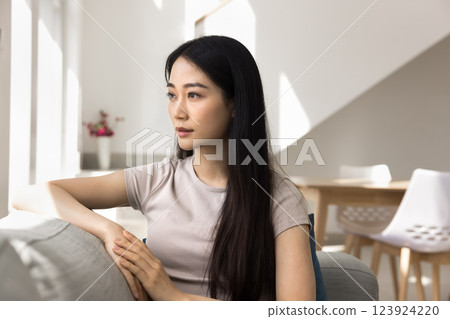 Serious Asian young woman sits thoughtfully on comfortable sofa 123924220