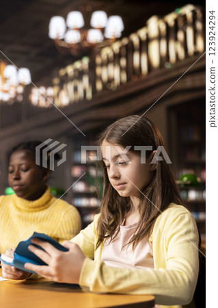 Young teenagers reading literary texts at the library as part of their curriculum, preparing for literature class lesson and enhancing their academic focus. Girls read the mandatory books titles. 123924294