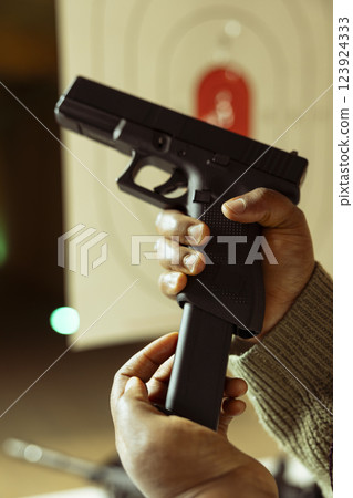 Close up of hands loading ammo in gun at indoor firing range with paper target in blurry background. Person doing shooting practice, learning how to responsibly empty pistol ammunition 123924333