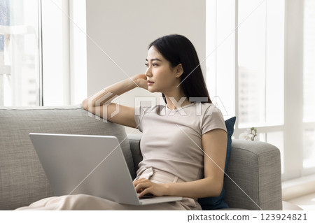 Woman gazing thoughtfully into distance seated on sofa with laptop Woman gazing thoughtfully into distance seated on sofa with laptop 123924821