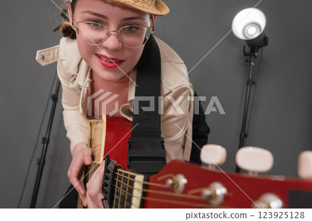 Cheerful female guitarist with glasses and grin on face is holding guitar and pointing it at camera. Playful expression musician is having fun, enjoying playing guitar during performance on stage 123925128