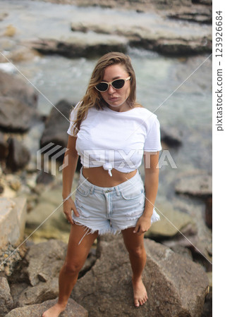A woman in a white crop top and shorts stands on a rock by water A woman in a white crop top and shorts stands on a rock by water 123926684