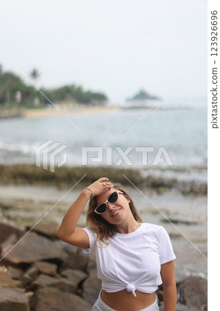 A woman in a white crop top and shorts stands on a rock by water A woman in a white crop top and shorts stands on a rock by water 123926696