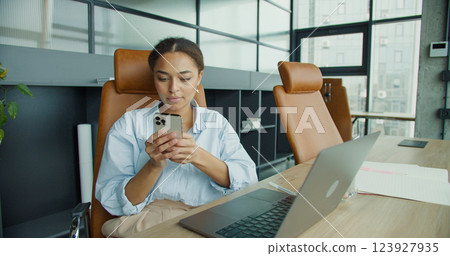 young woman checking her smartphone while working on a laptop in a modern office, sitting at a desk with notebooks, a glass of water, and stylish leather chairs 123927935
