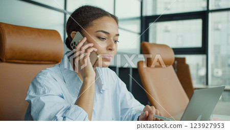 A young woman multitasking in a modern office, talking on the phone while holding a pen, looking focused and engaged at her workspace with a laptop A young woman multitasking in a modern office, talking on the phone while holding a pen, looking focused and engaged at her workspace with a laptop 123927953