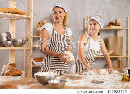 Two women working at bakery, kneading dough and rolling out dough into sausages and shaping baguettes on countertop in kitchen Two women working at bakery, kneading dough and rolling out dough into sausages and shaping baguettes on countertop in kitchen 123928723