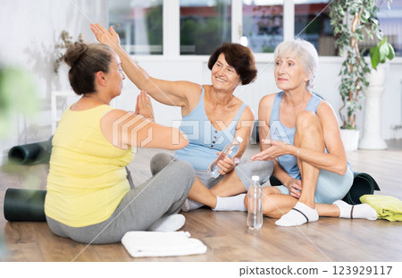 Mature women talking to each other sitting on the floor in fitness studio Mature women talking to each other sitting on the floor in fitness studio 123929117