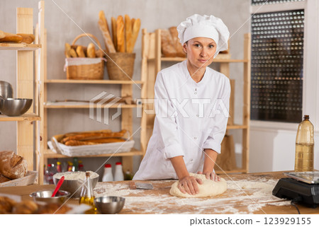 Experienced young woman in white uniform working in bakery kitchen, kneading raw dough 123929155