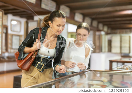 Young woman with teen daughter visitors stands near horizontal glass display case Young woman with teen daughter visitors stands near horizontal glass display case 123929503