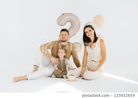 Caucasian young family of three, a man, a woman and a baby, are posing for a picture in front of a birthday cake and balloons Caucasian young family of three, a man, a woman and a baby, are posing for a picture in front of a birthday cake and balloons 123932659