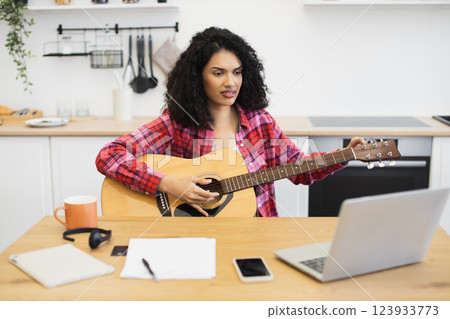 Young adult woman with long curly hair in casual attire playing guitar during an online lesson in kitchen. Technology and learning concept featuring acoustic guitar, laptop, notes. Young adult woman with long curly hair in casual attire playing guitar during an online lesson in kitchen. Technology and learning concept featuring acoustic guitar, laptop, notes. 123933773