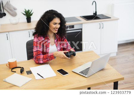 Young African American woman wearing plaid shirt enjoying video game while interacting online. Her setup includes gaming controller, laptop, and casual work items in a bright, organized kitchen. Young African American woman wearing plaid shirt enjoying video game while interacting online. Her setup includes gaming controller, laptop, and casual work items in a bright, organized kitchen. 123933779