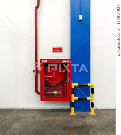 A fire hose reel is mounted on a white wall, accompanied by a bright red sign. A yellow and black safety barrier line the area, ensuring visibility and safety. 123934920