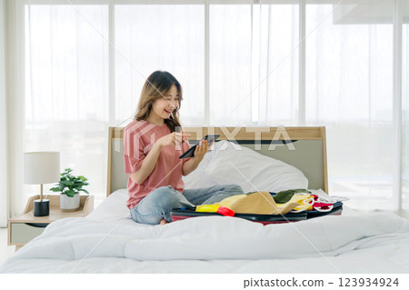 A young woman sit on a bed, smiling and engaged with her tablet computer while packing a suitcase. Various colorful cloth and accessory are spread around her in bedroom. 123934924