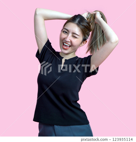 A young woman smile and wink while styling her hair into a ponytail. She wear a black t-shirt and is set against a soft pink background, creating a lively atmosphere. A young woman smile and wink while styling her hair into a ponytail. She wear a black t-shirt and is set against a soft pink background, creating a lively atmosphere. 123935114