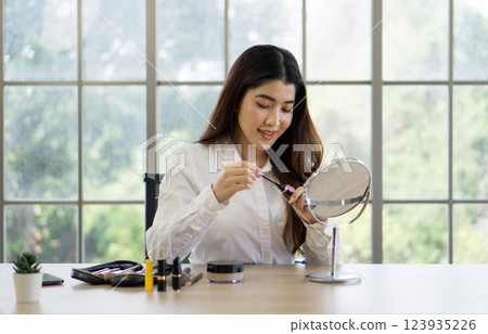 Young asian woman focused on applying mascara. There are  collection of makeup items and brushes in front of her as well as a makeup palette 123935226