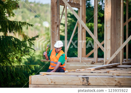 Carpenter constructing two-story wooden frame house near the forest. Bearded man wearing glasses hammering nails into structure. Concept of modern ecological construction. Carpenter constructing two-story wooden frame house near the forest. Bearded man wearing glasses hammering nails into structure. Concept of modern ecological construction. 123935922