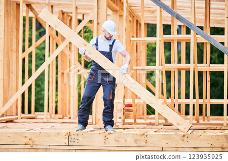 Carpenter constructing wooden-framed house. Man holding large joist and dressed in work clothes and helmet. Concept of modern and environmentally-friendly construction. Carpenter constructing wooden-framed house. Man holding large joist and dressed in work clothes and helmet. Concept of modern and environmentally-friendly construction. 123935925