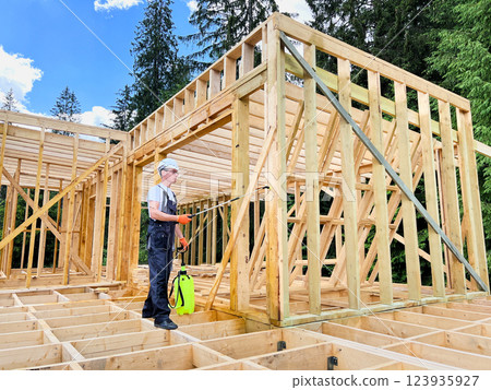 Laborer constructing wooden frame house near forest. Man treating woods, applying fire retardant using sprayer, while dressed in protective suit, helmet. Concept of modern eco-friendly construction. Laborer constructing wooden frame house near forest. Man treating woods, applying fire retardant using sprayer, while dressed in protective suit, helmet. Concept of modern eco-friendly construction. 123935927