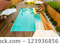 Elegant backyard with turquoise swimming pool, wooden deck, umbrellas, and lounge chairs in a sunny setting 123936856