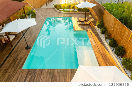 Elegant backyard with turquoise swimming pool, wooden deck, umbrellas, and lounge chairs in a sunny setting Elegant backyard with turquoise swimming pool, wooden deck, umbrellas, and lounge chairs in a sunny setting 123936856