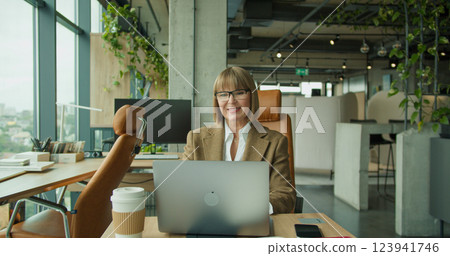 A senior professional woman working on a laptop in a modern office setting with large windows and lush greenery, symbolizing efficiency and sophistication 123941746