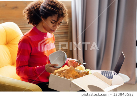 An African unpacks a cardboard box in the living room of the house. A charming black girl is outraged by a broken plate. An African unpacks a cardboard box in the living room of the house. A charming black girl is outraged by a broken plate. 123943582