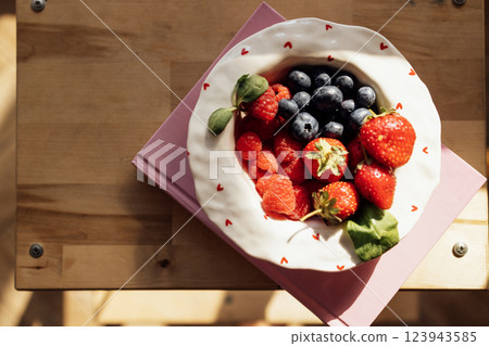 A cute porcelain plate with berries and a book on a wooden table. A healthy morning snack. Strawberries, raspberries, blueberries and basil leaves for breakfast. A cute porcelain plate with berries and a book on a wooden table. A healthy morning snack. Strawberries, raspberries, blueberries and basil leaves for breakfast. 123943585